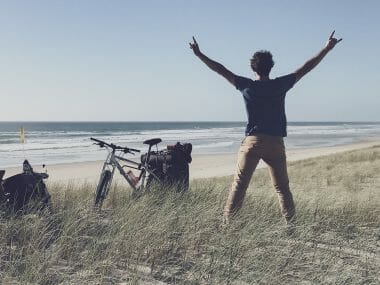 un homme lève les bras devant une plage et deux vélos