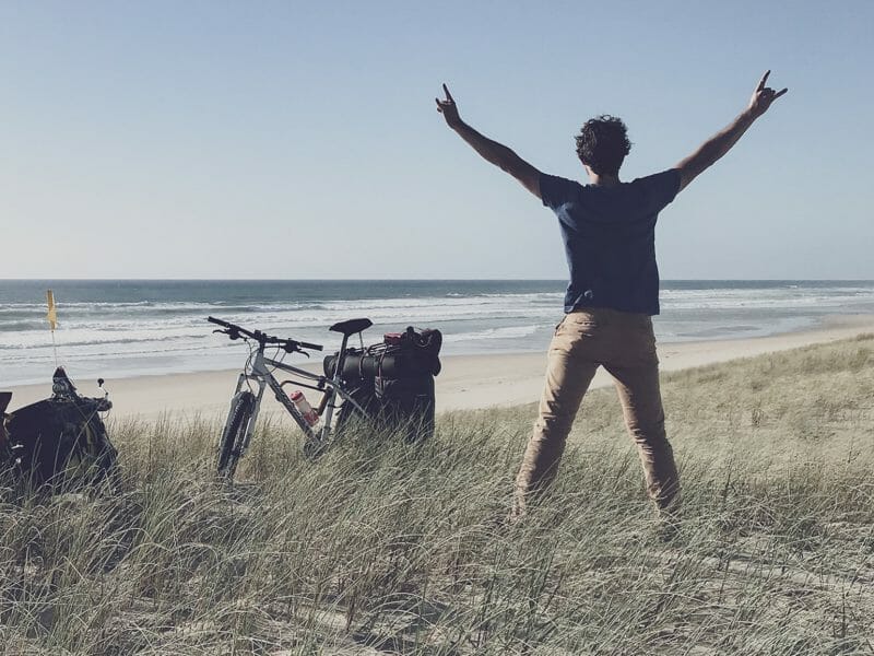 un homme lève les bras devant une plage et deux vélos