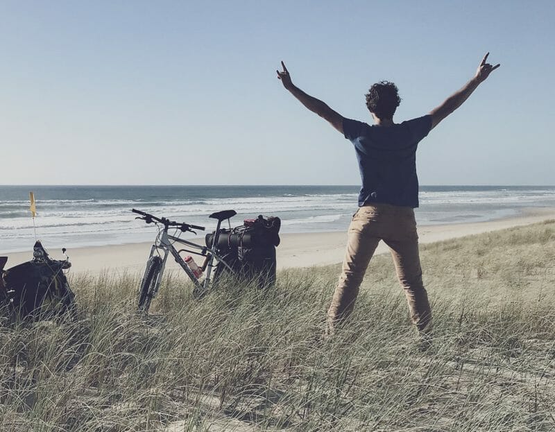 un homme lève les bras devant une plage et deux vélos