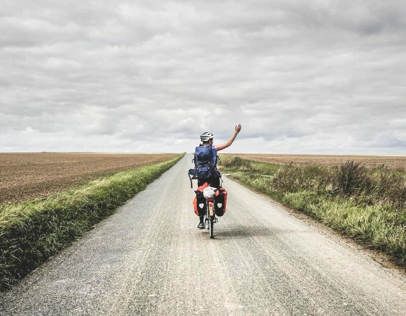 une femme de dos lève le bras en faisant du vélo sur une route de campagne